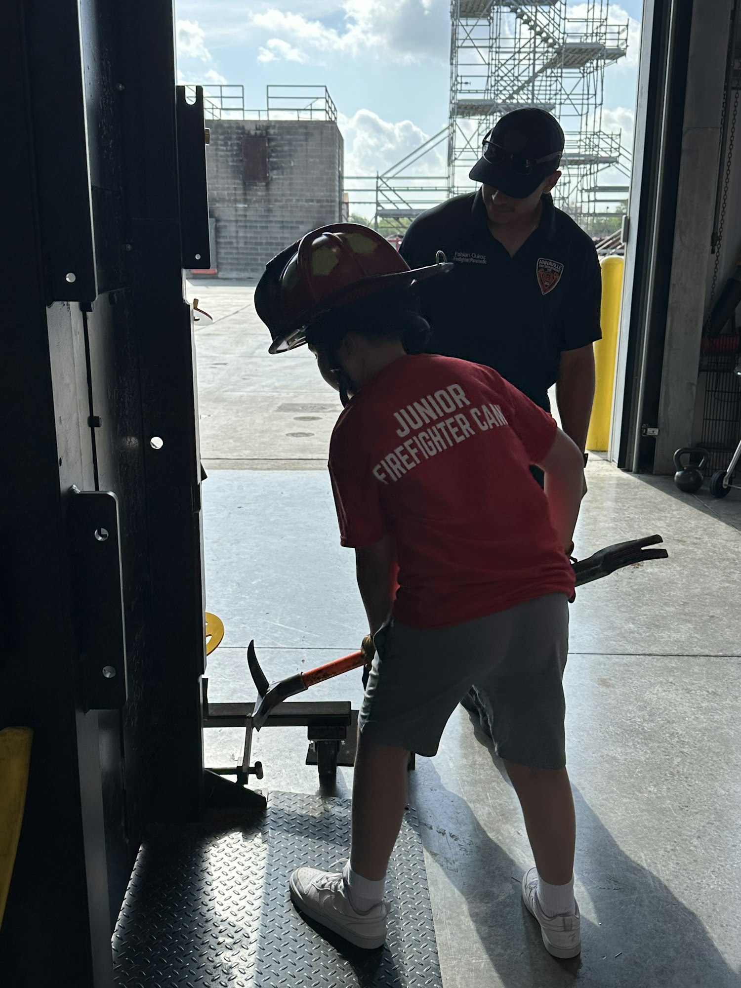 A child in a "Junior Firefighter Camp" shirt interacts with an adult in a workshop or training area.