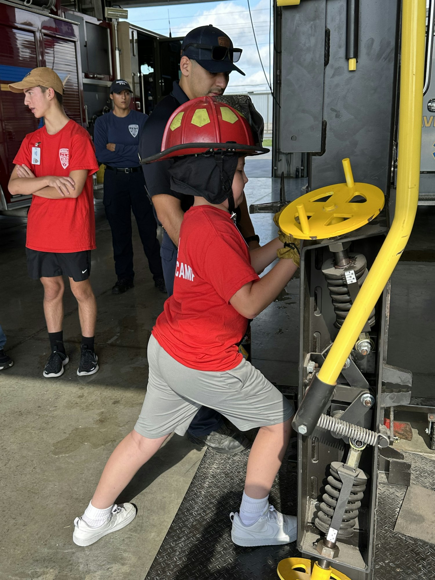 A person in a red shirt operates equipment, assisted by two others in a fire station setting.