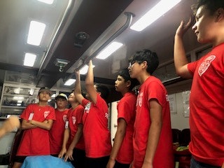 A group of people in red shirts are standing together inside a room or vehicle, some raising their hands.