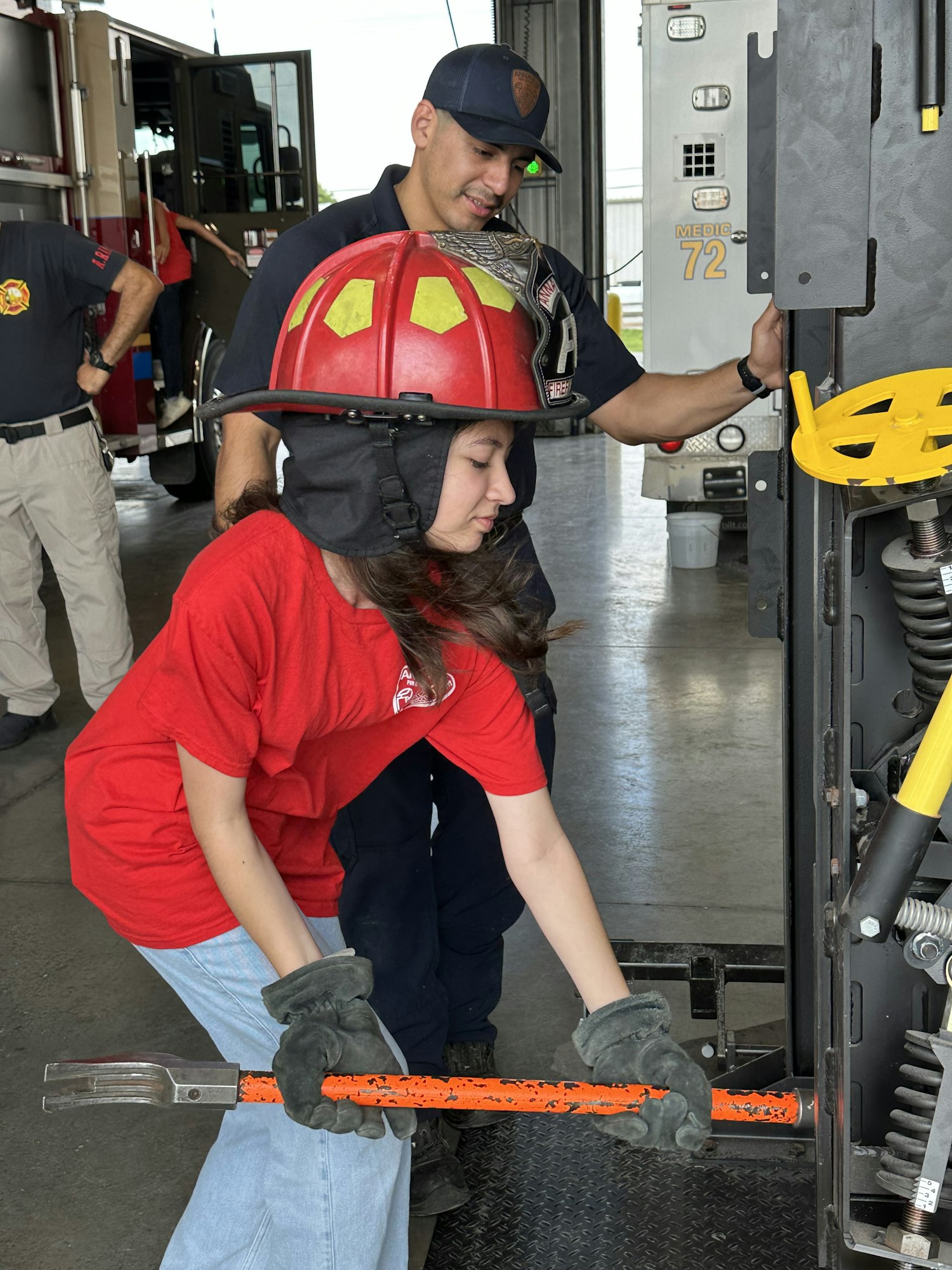 A person in a red helmet and shirt uses a tool with guidance from a firefighter in a station.