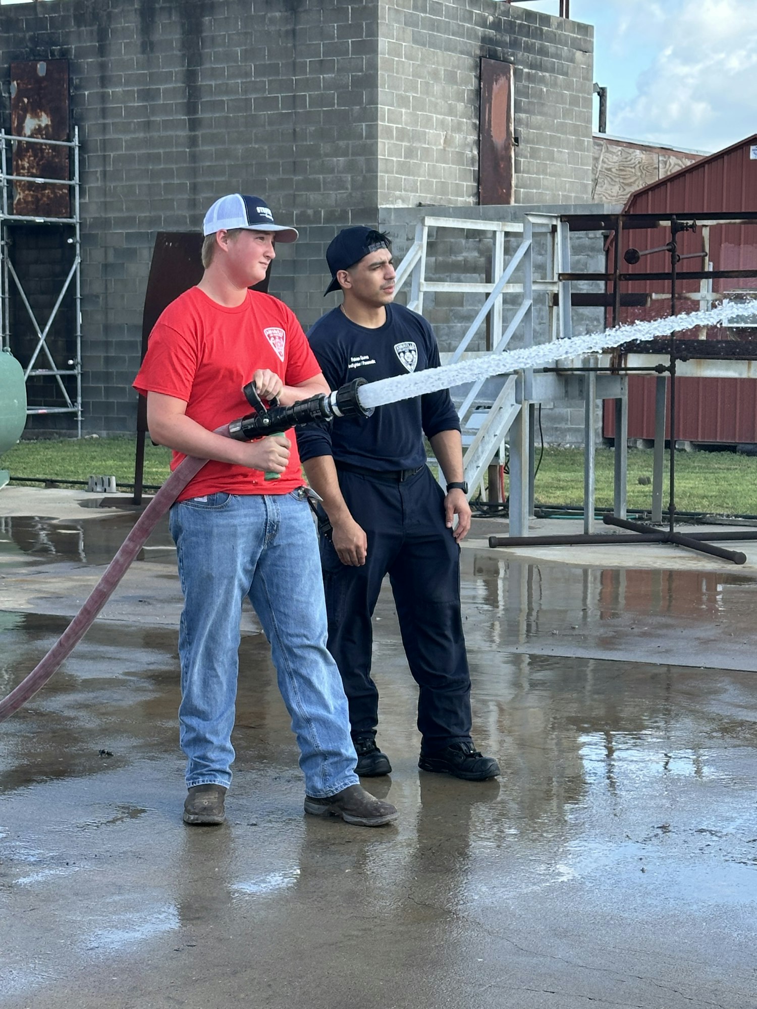 Two people are spraying water from a fire hose outside near a brick building.