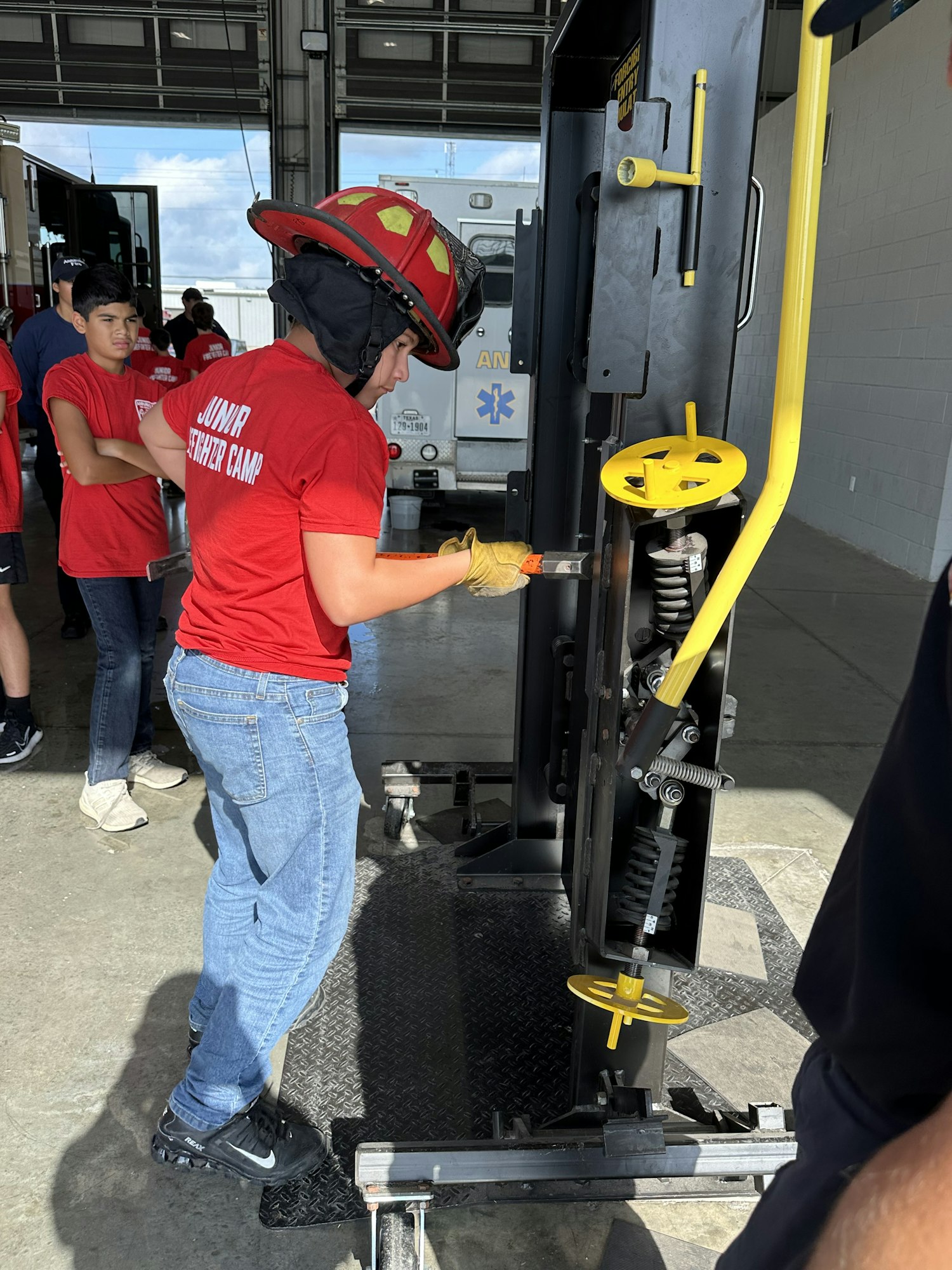 A person in a firefighter helmet uses a tool on a metal structure, with others observing in the background.