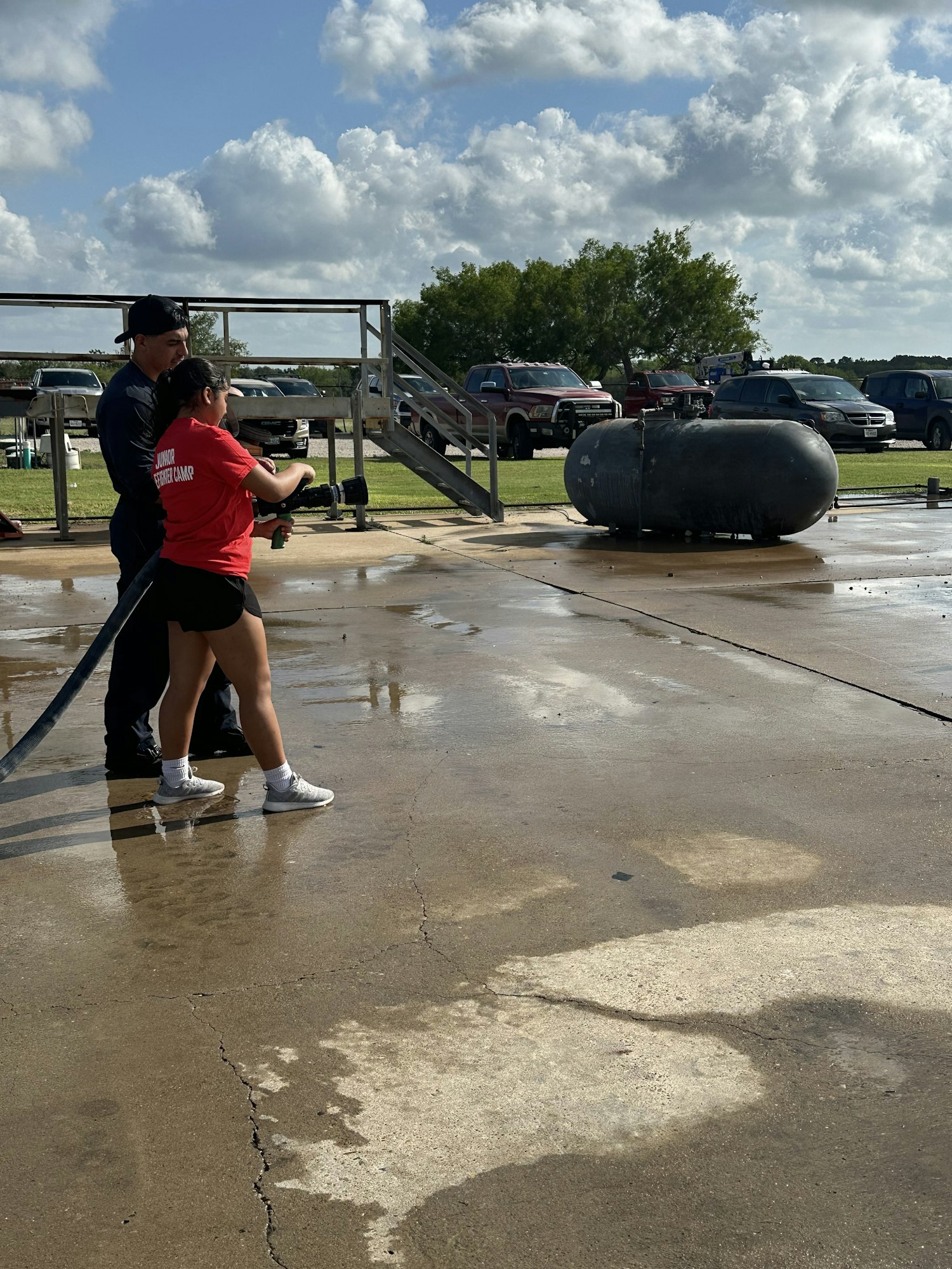 Two people, one in red, using a hose on a wet pavement with parked cars and trees in the background.