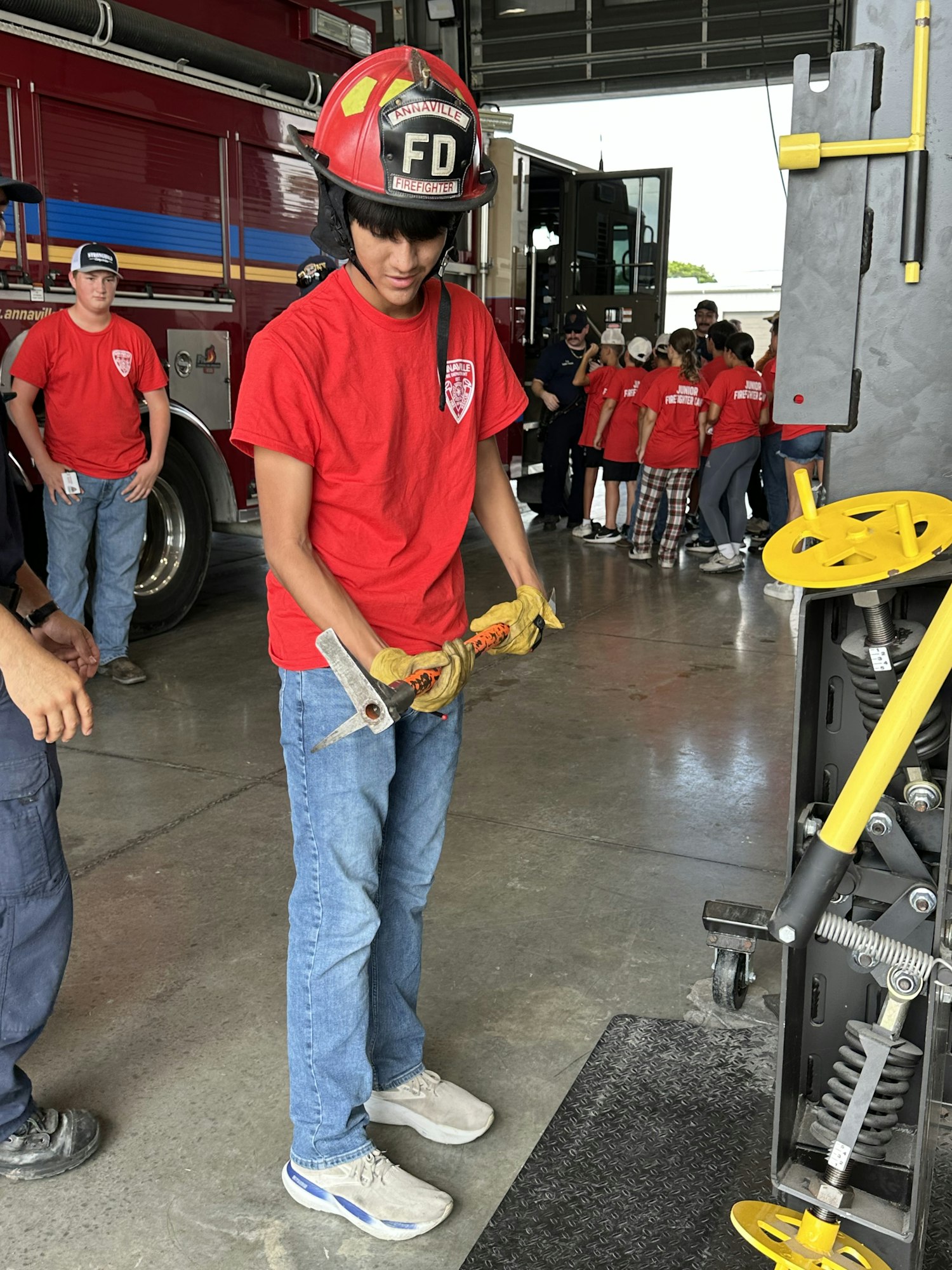 A person in a red shirt and firefighter helmet is handling equipment in a garage setting. A group watches in the background.