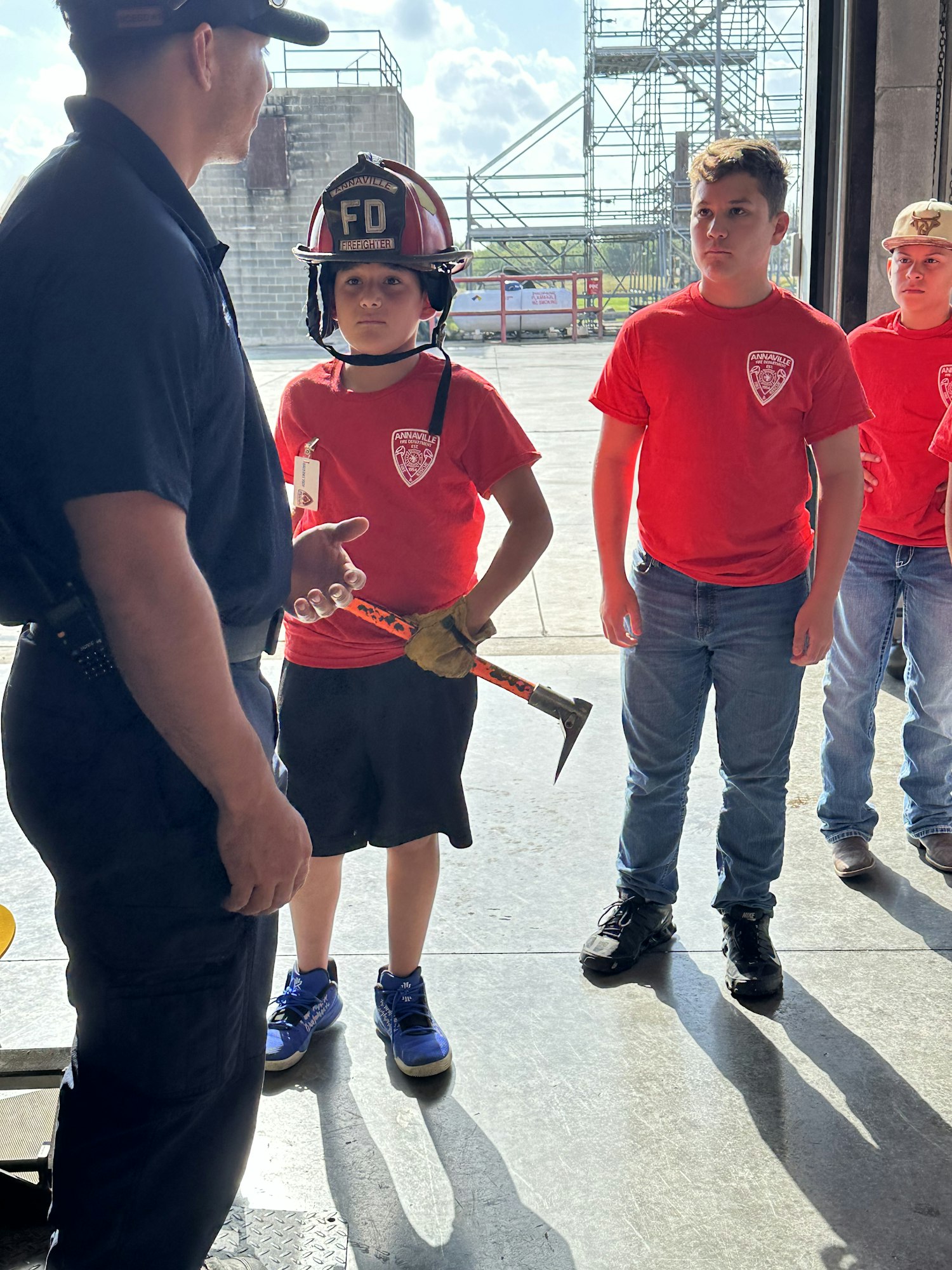 A child in a fire helmet holds a tool, talking to a person in uniform, with others in red shirts nearby.