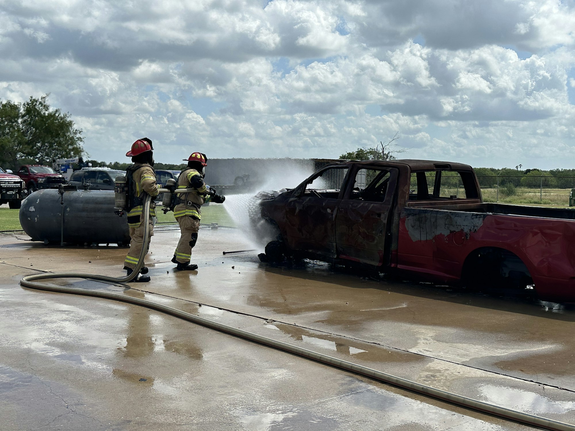 Firefighters extinguishing a fire on a burned-out truck using a hose, with a propane tank nearby.