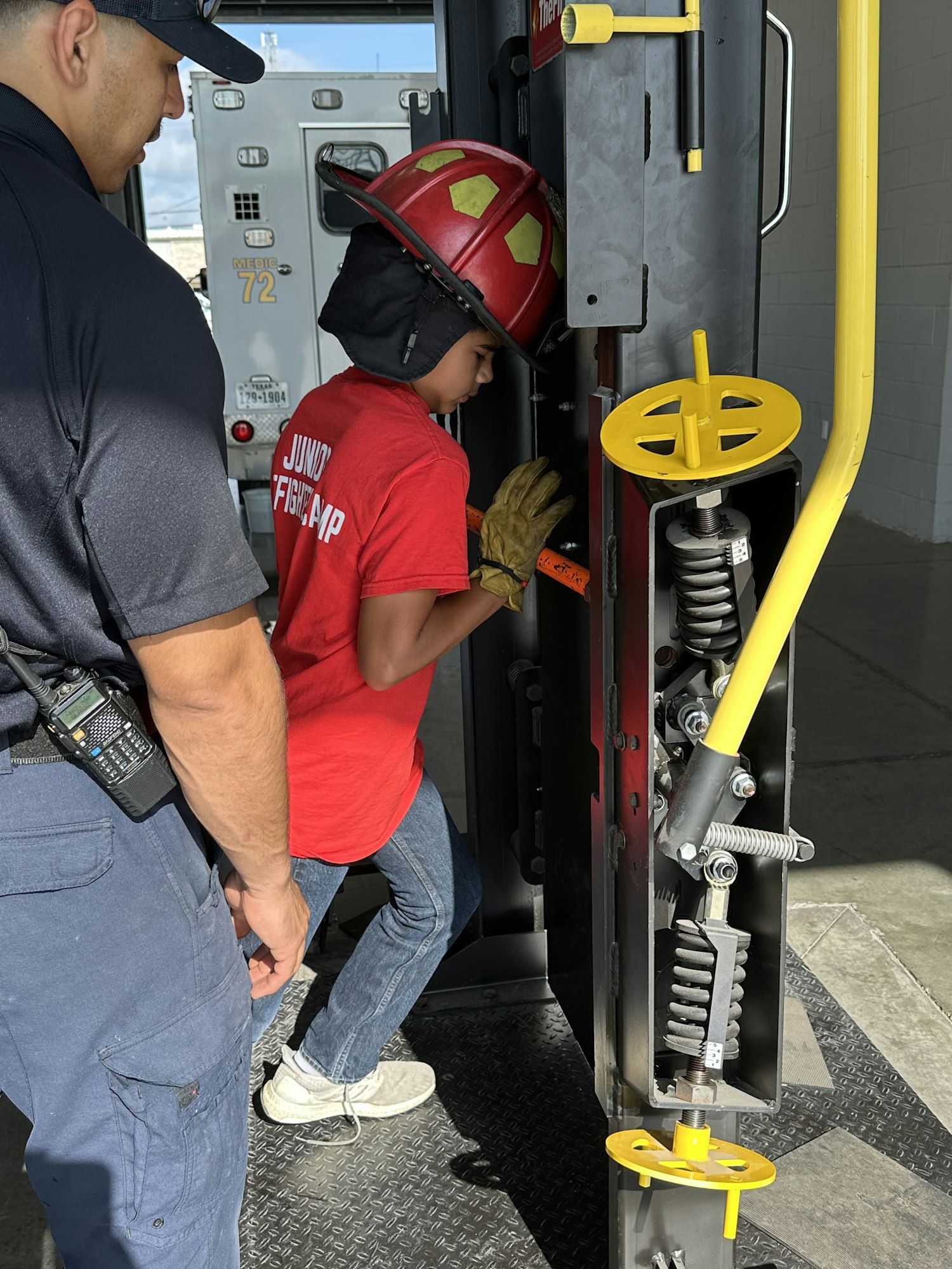 A child in a red helmet and shirt labeled "Junior Firefighter" interacts with fire equipment, supervised by an adult.