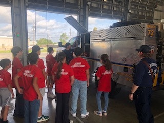 A group of children in red shirts is listening to a firefighter next to a fire truck inside a fire station.