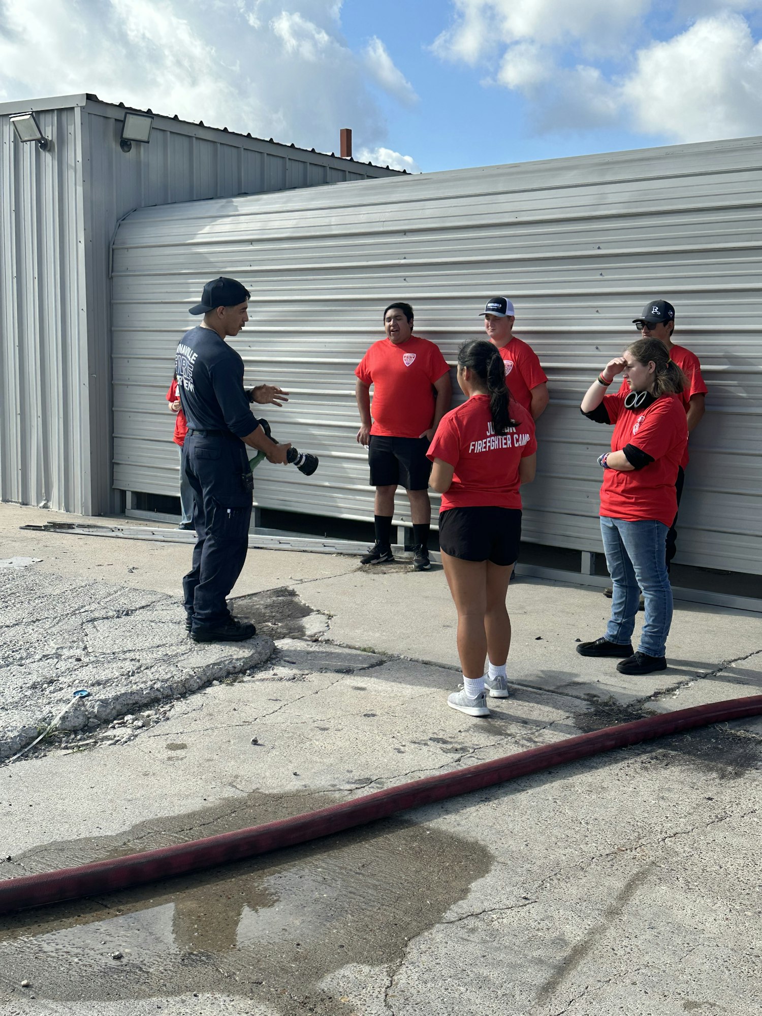 A group of people, some in red shirts labeled "FIREMEN," standing near a metal building and talking, with a hose on the ground.