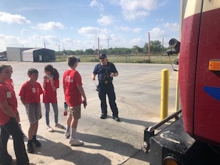 A group of people in red shirts stand outside near a large vehicle, possibly receiving a demonstration or instructions from a uniformed person.