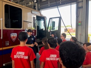 A firefighter is giving a tour or lesson to a group of kids in "Junior Firefighter Camp" shirts near a fire truck.