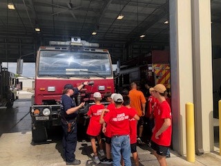 A group of people in red shirts stand around a large fire truck at a fire station.