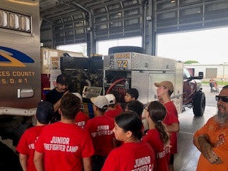 A group of kids in red "Junior Firefighter Camp" shirts are gathered in a garage near fire trucks, with an adult explaining something.