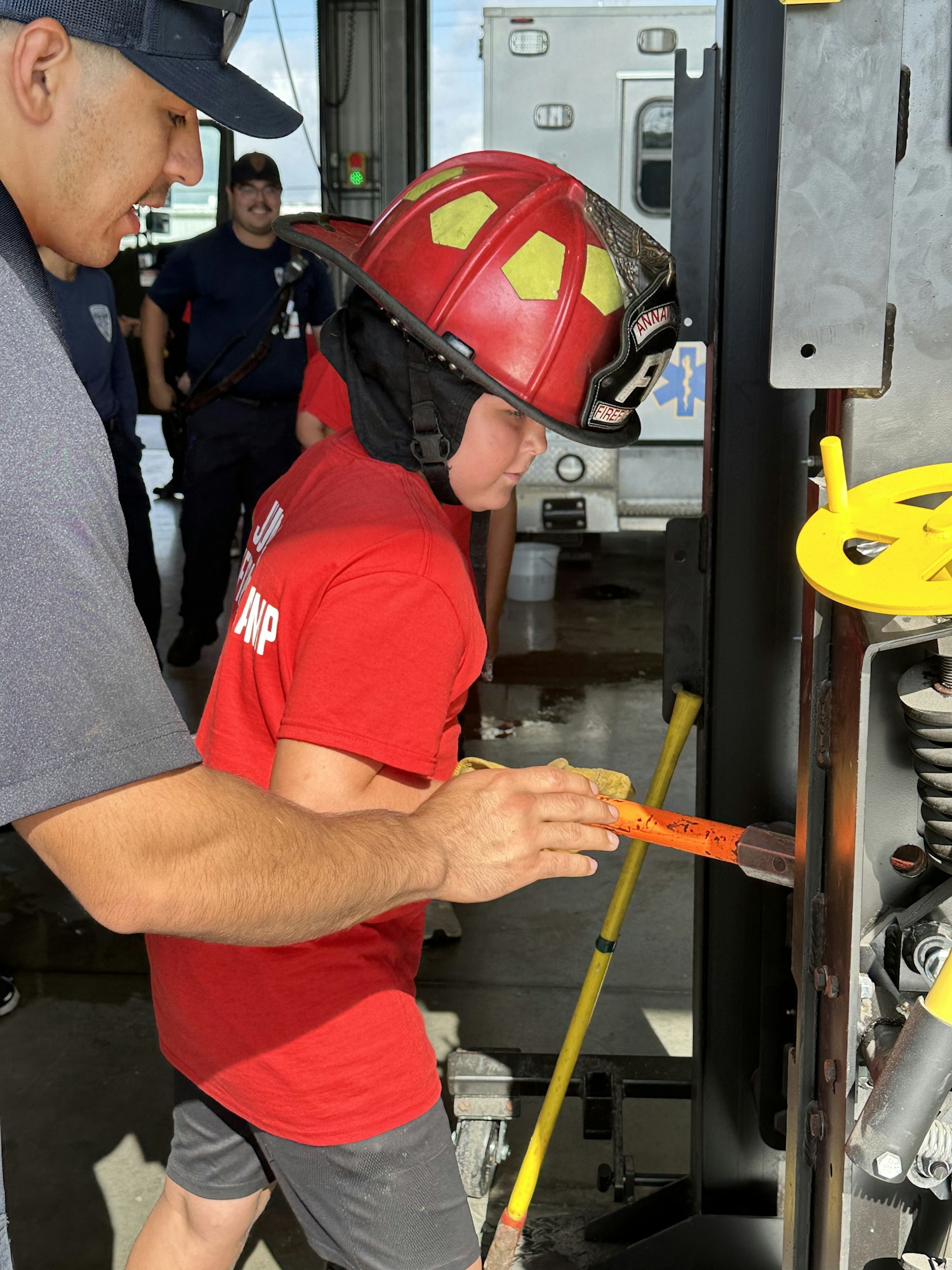 A person in a red helmet is engaged in an activity with an orange tool, supervised by others, possibly at a fire station.