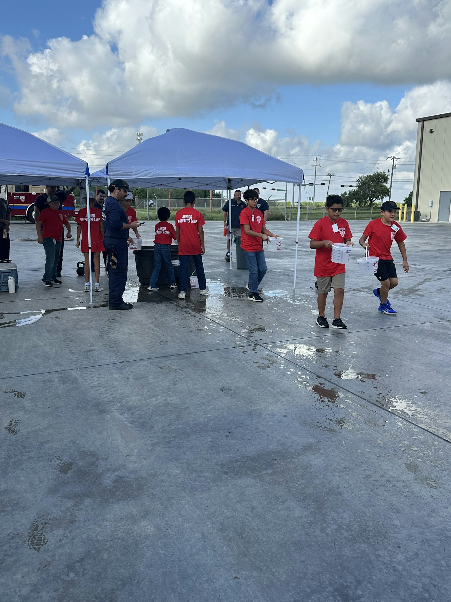 People in red shirts stand under a white tent on wet pavement, possibly at an event with chairs and signage in the background.