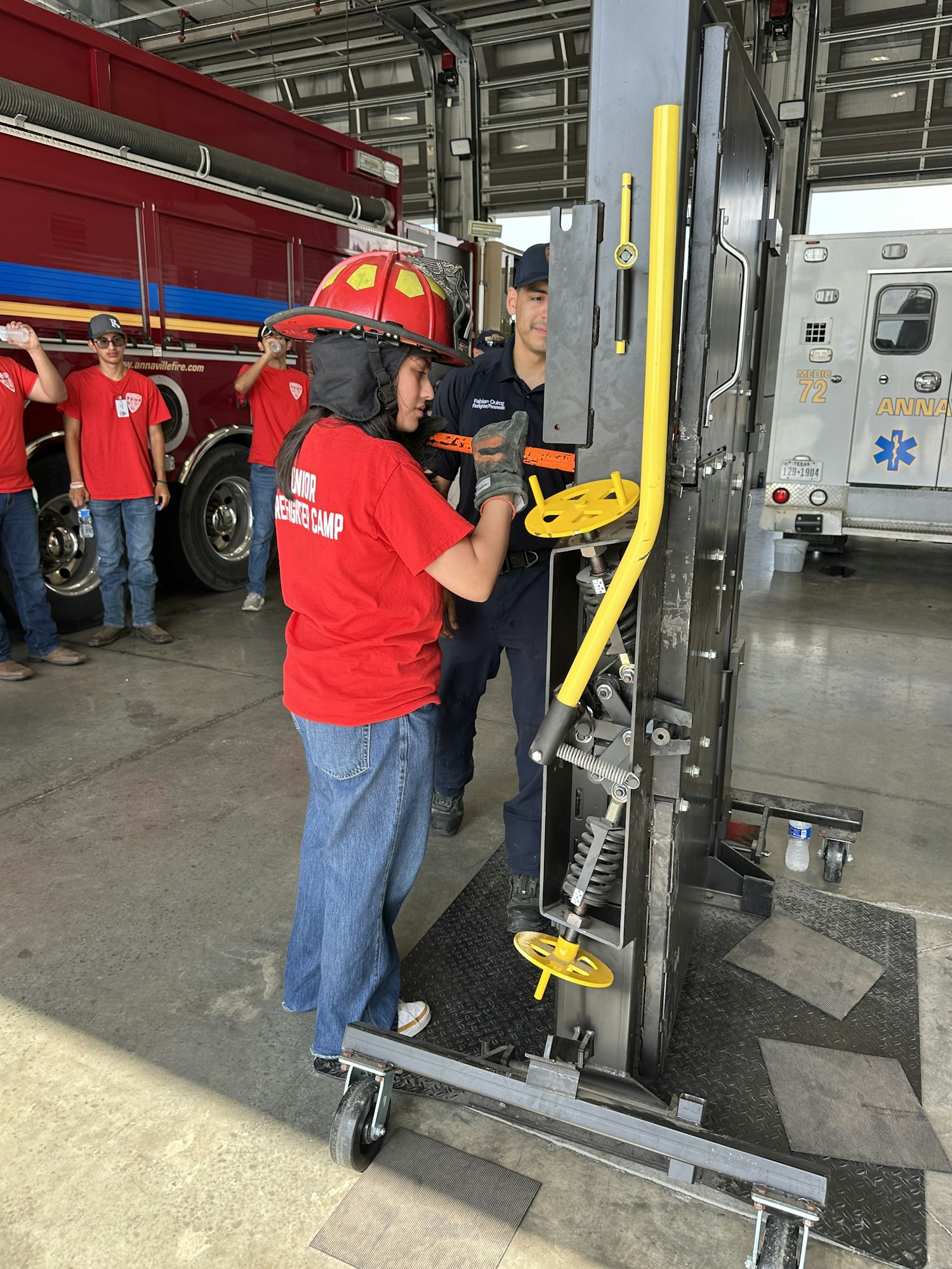 A firefighter instructs a person in a red shirt and helmet using equipment near a fire truck and ambulance.
