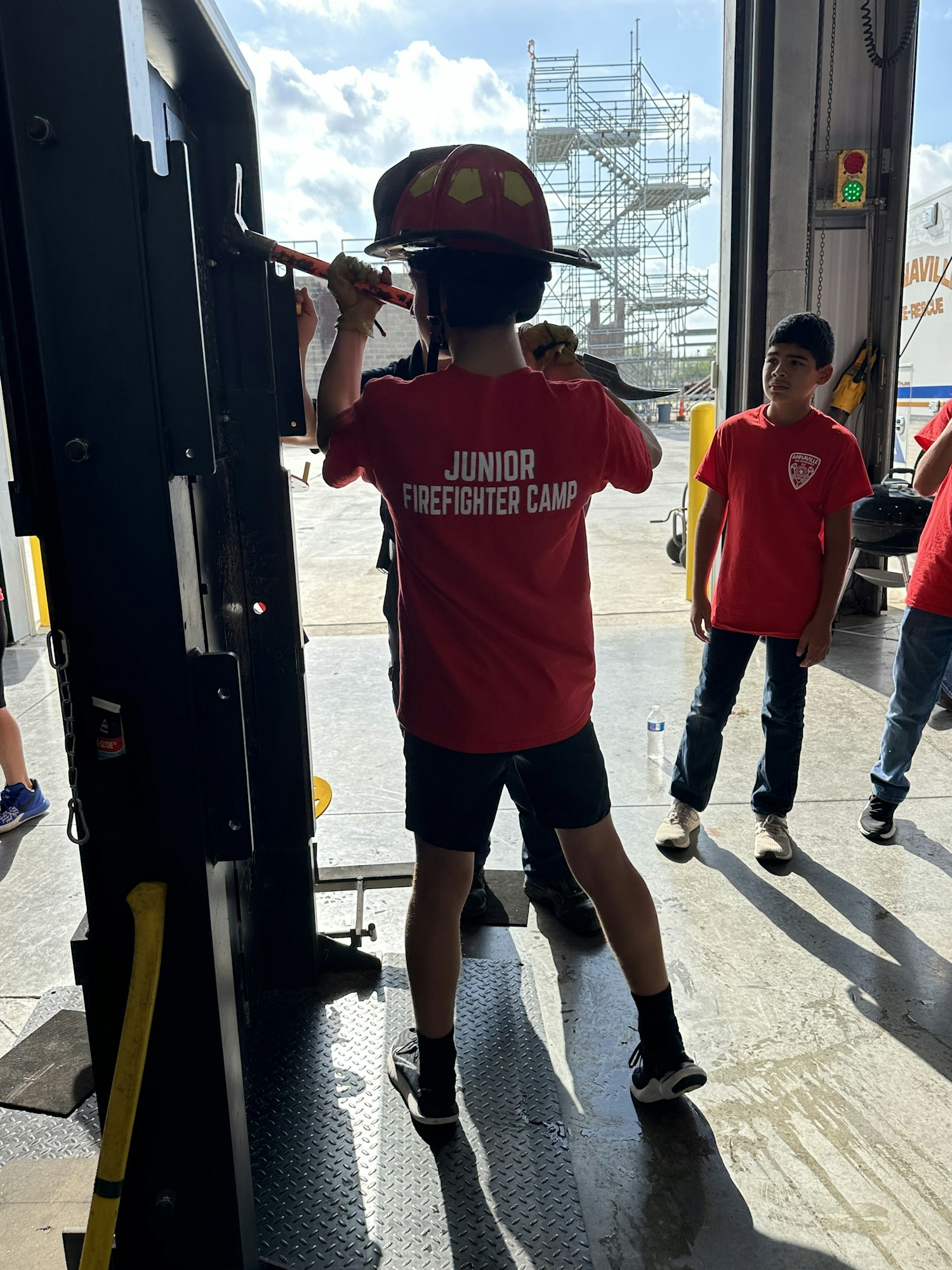Child in a "Junior Firefighter Camp" shirt using a tool inside a garage, with people watching.