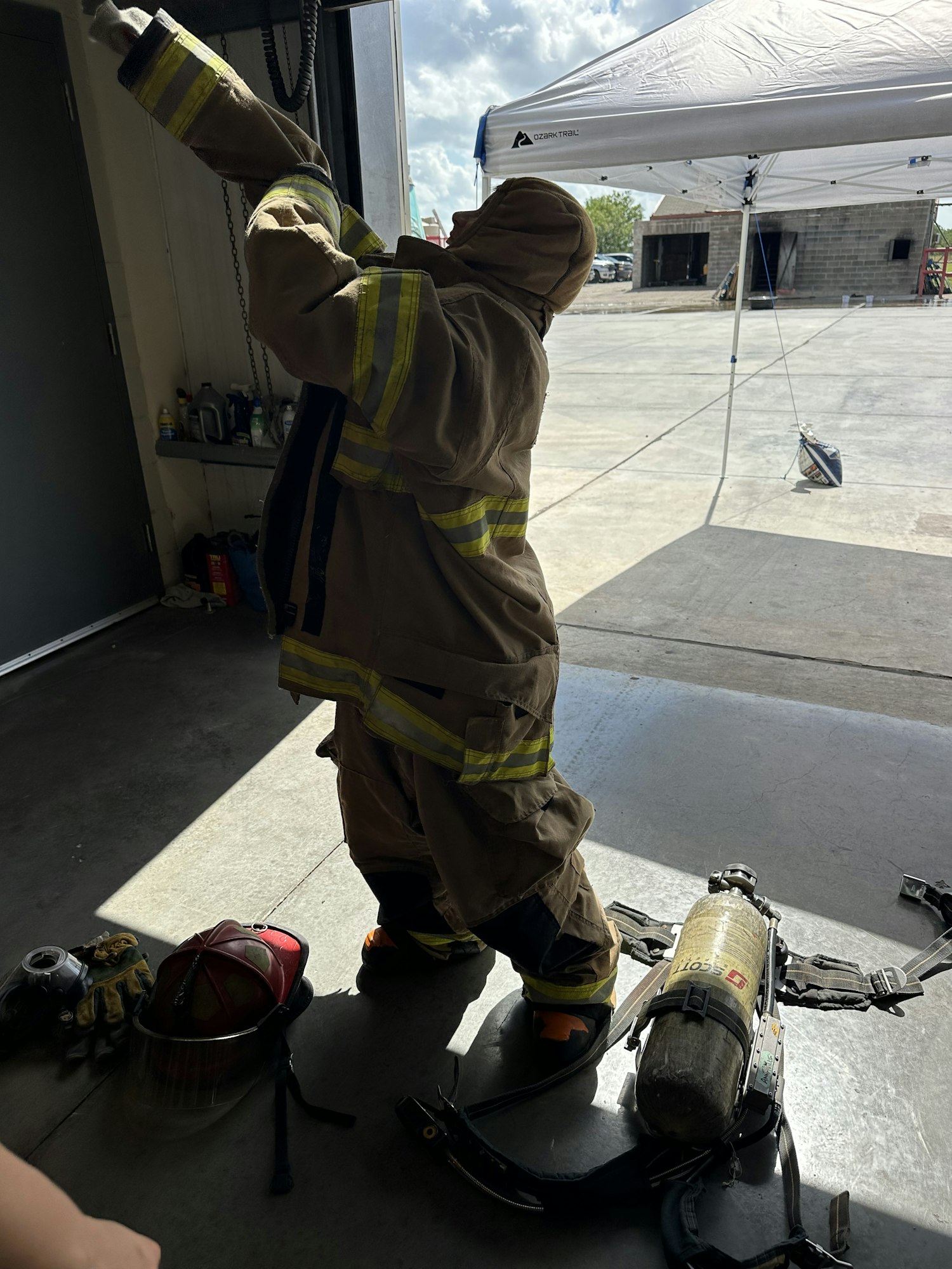 A firefighter in full gear, including helmet and air tank, preparing for duty in a garage or station.