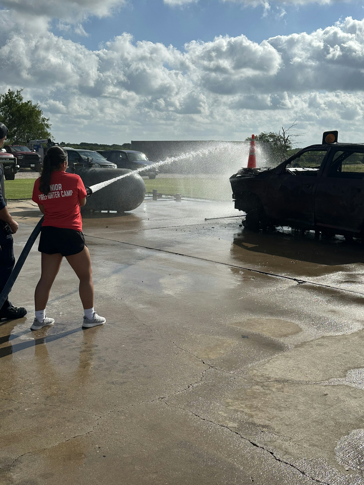 Person in red shirt spraying water from a hose at a burnt vehicle, with cloudy sky and wet ground.