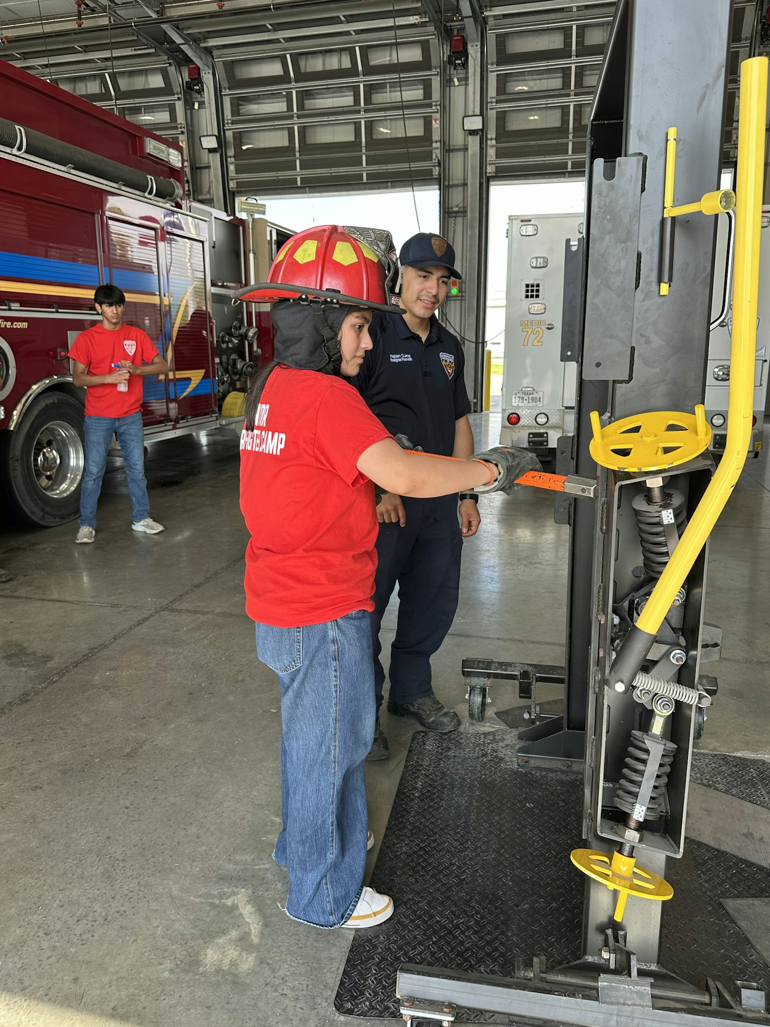 People in a fire station, one in a red helmet adjusting equipment. A fire truck is in the background.