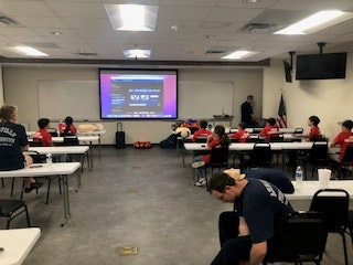 Classroom setting with people in red shirts watching a presentation on a screen. Desks and CPR practice mannequins are visible.
