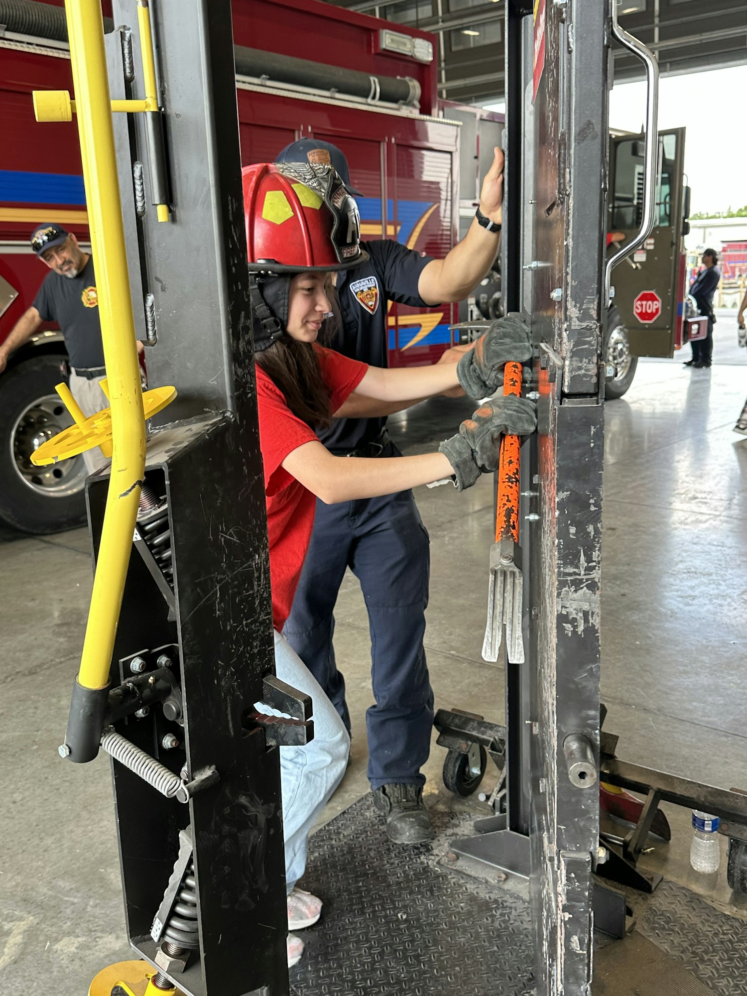 Two people in firefighter gear engage with a large metallic door or equipment, possibly a training exercise at a fire station.