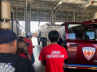 Group of kids in a fire station, one wearing a "Junior Firefighter Camp" shirt, near a red truck with "Annaville Fire Department" logo.