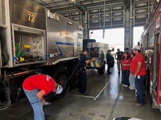 People working in a garage with a fire truck and equipment, some wearing red shirts, engaged in various tasks.