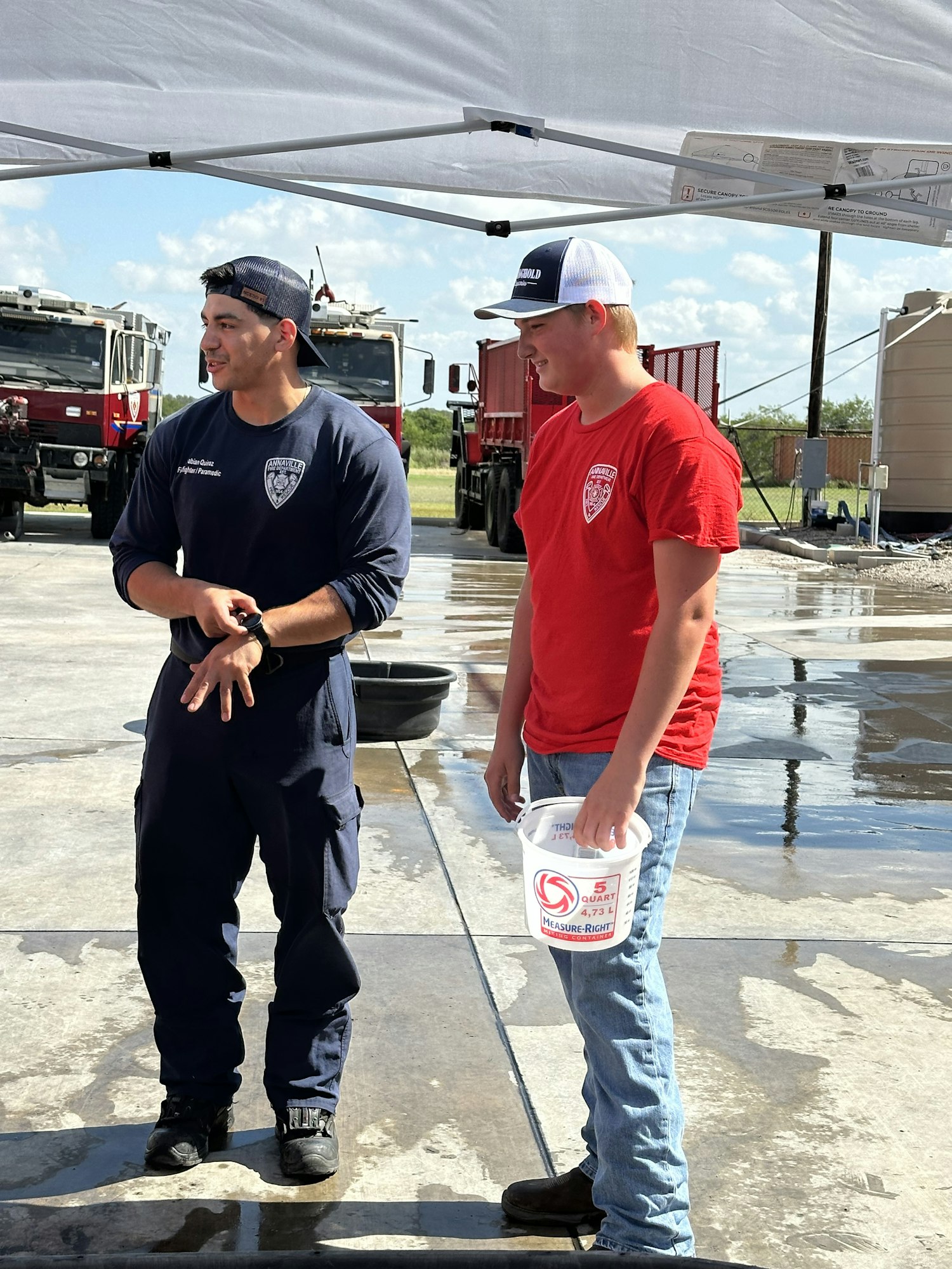 Two people in uniforms stand outdoors, one holding a bucket, with fire trucks and a tent visible.