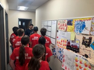 A group of kids in red "Junior Firefighter Camp" shirts in a hallway with a board and children's artwork on the wall.