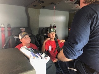 Two kids in red shirts and helmets sit inside a vehicle, facing an adult in a blue uniform.