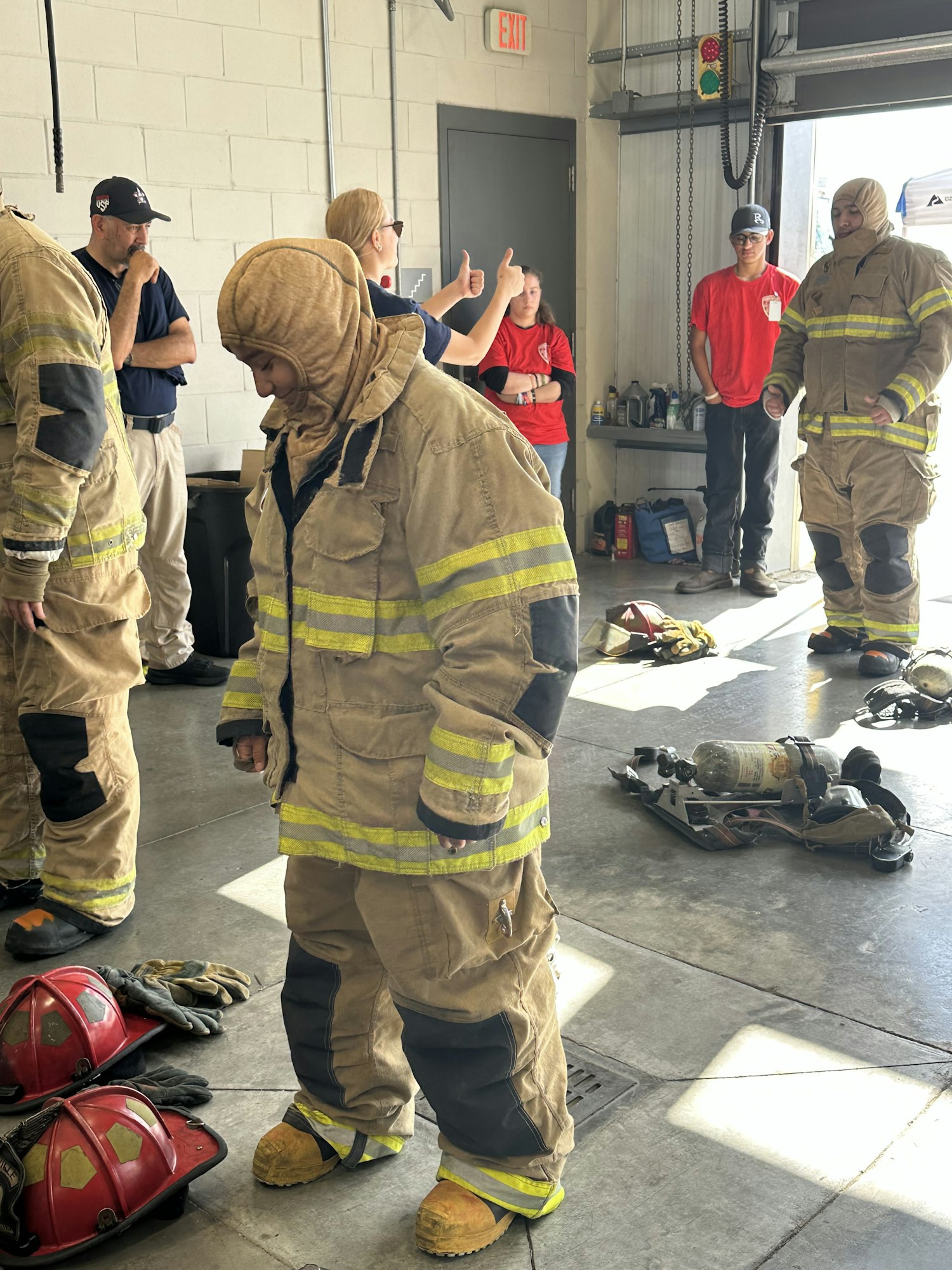 Firefighters in gear stand with others in a station, equipment on the floor.