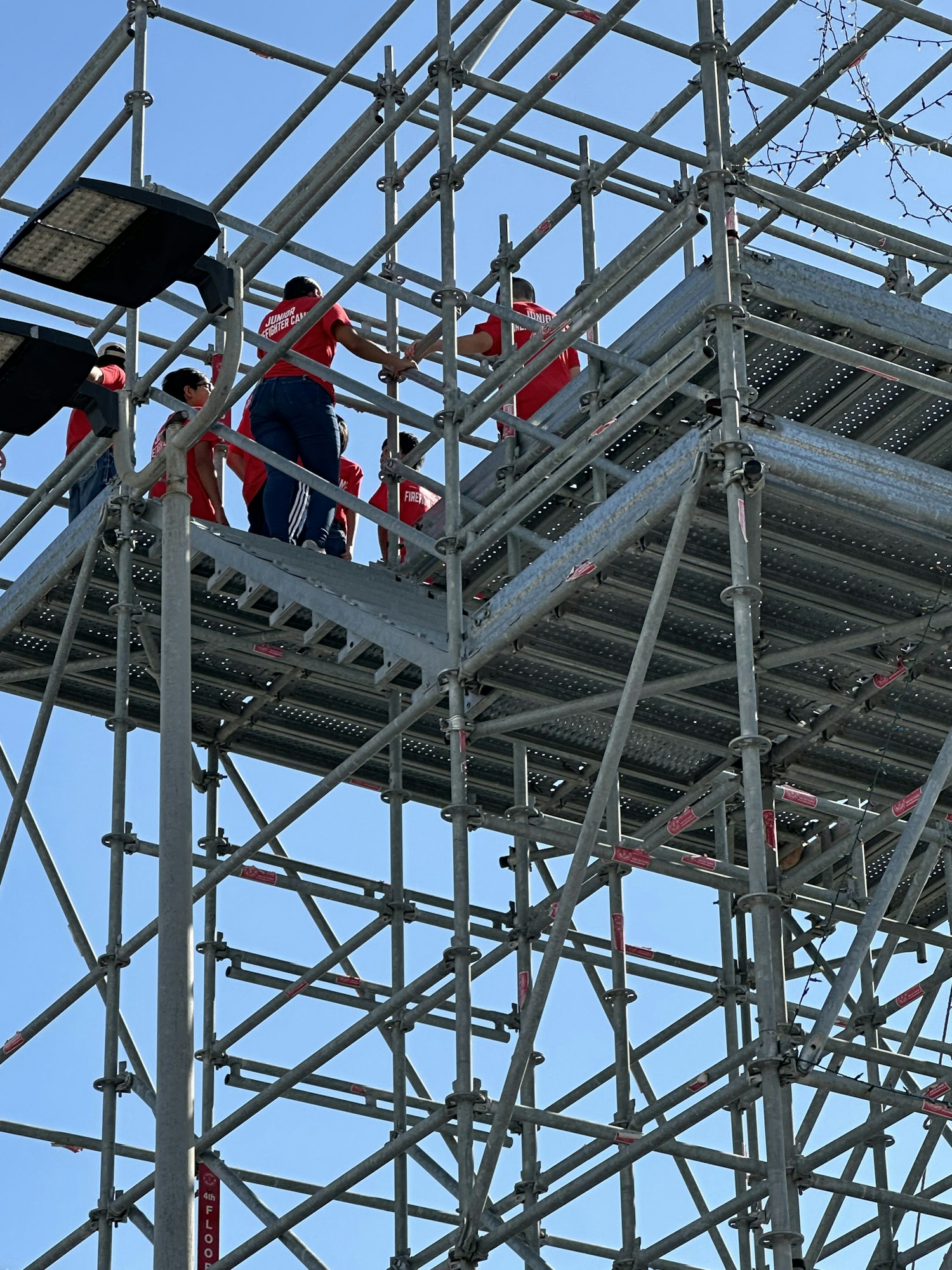 Workers in red uniforms climb scaffolding framed against a clear blue sky.