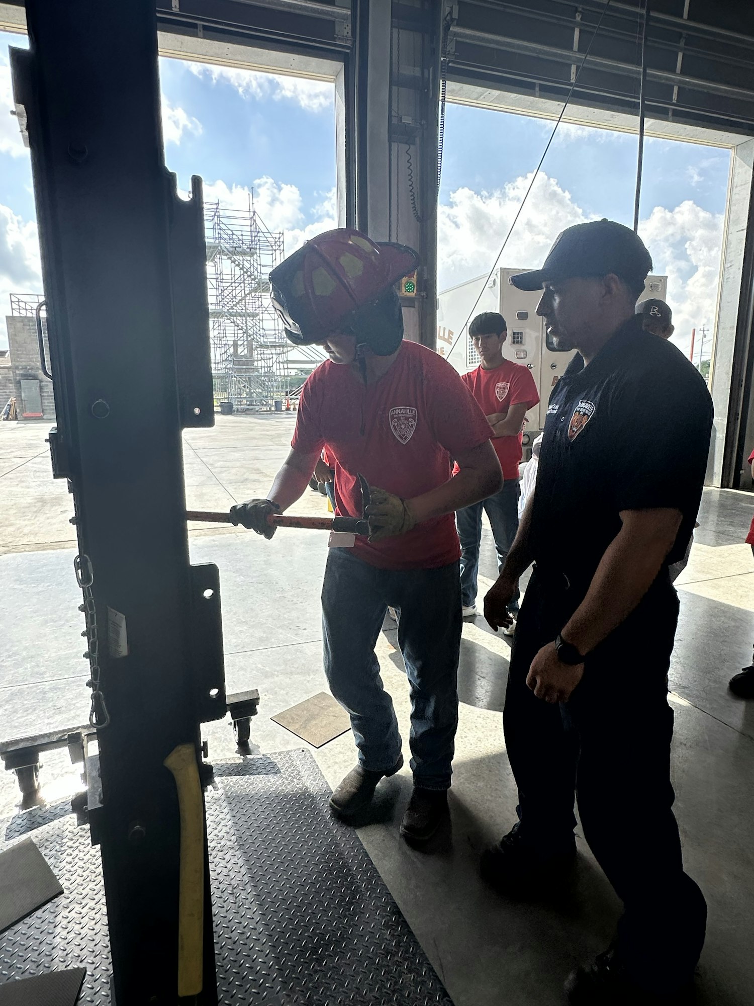 A firefighter in a helmet holds a tool inside a garage, with others in red shirts nearby.
