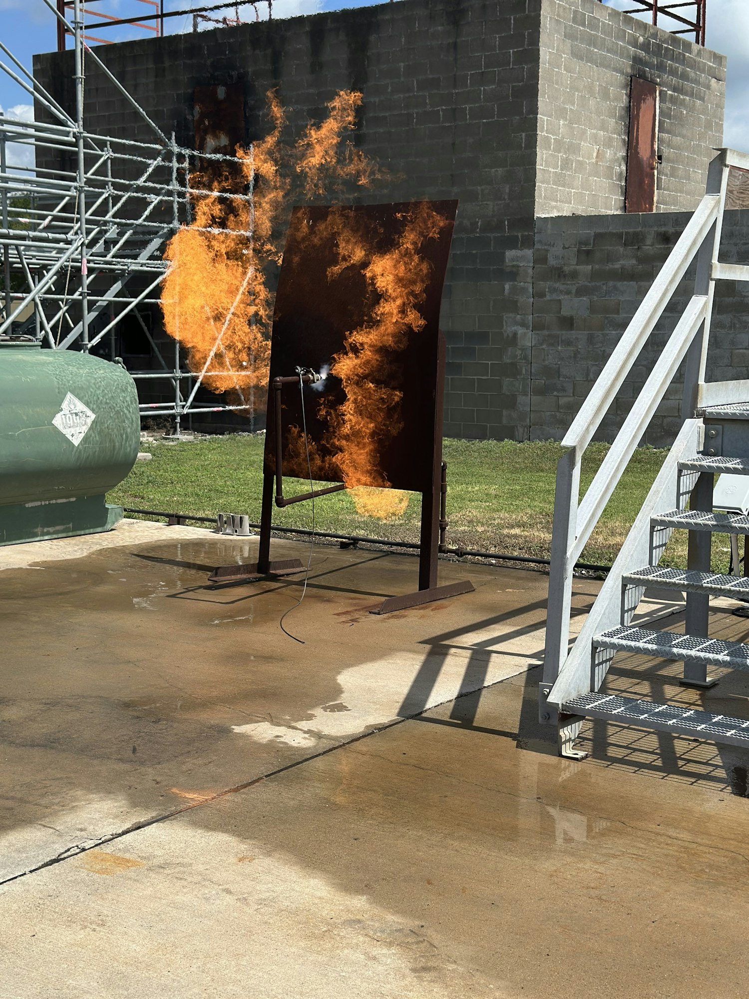A fire behind a metal structure on a concrete surface, with a building and scaffolding nearby.