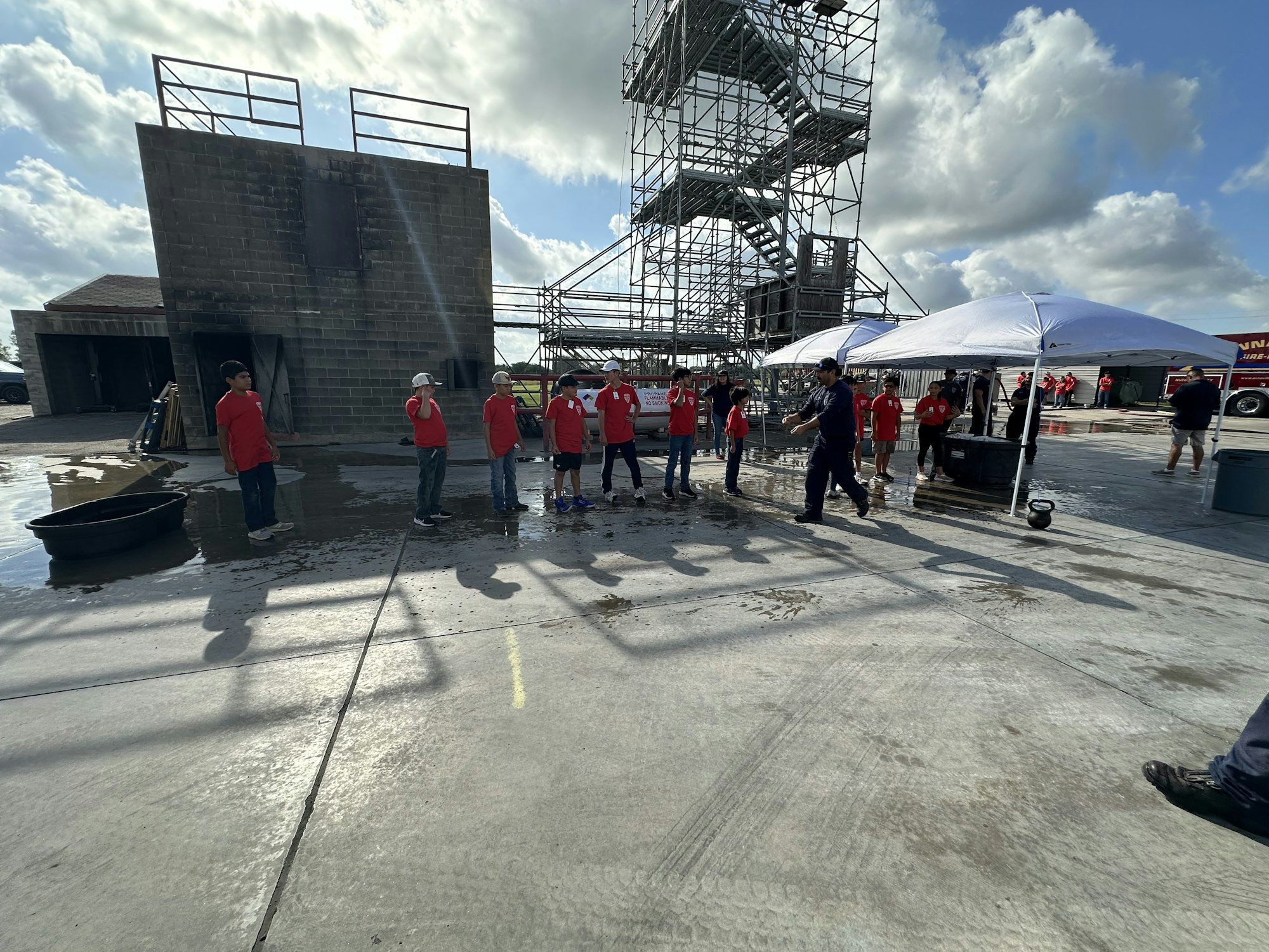 Group of people in red shirts standing near a building and scaffolding, with umbrellas and a sunny sky.