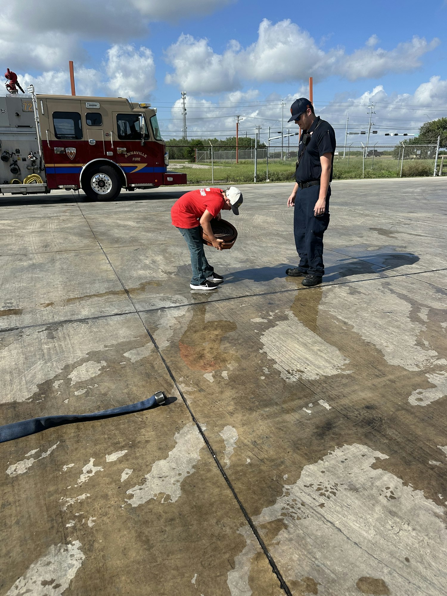 Two people are by a fire truck on a wet concrete surface, one holding a coiled hose.