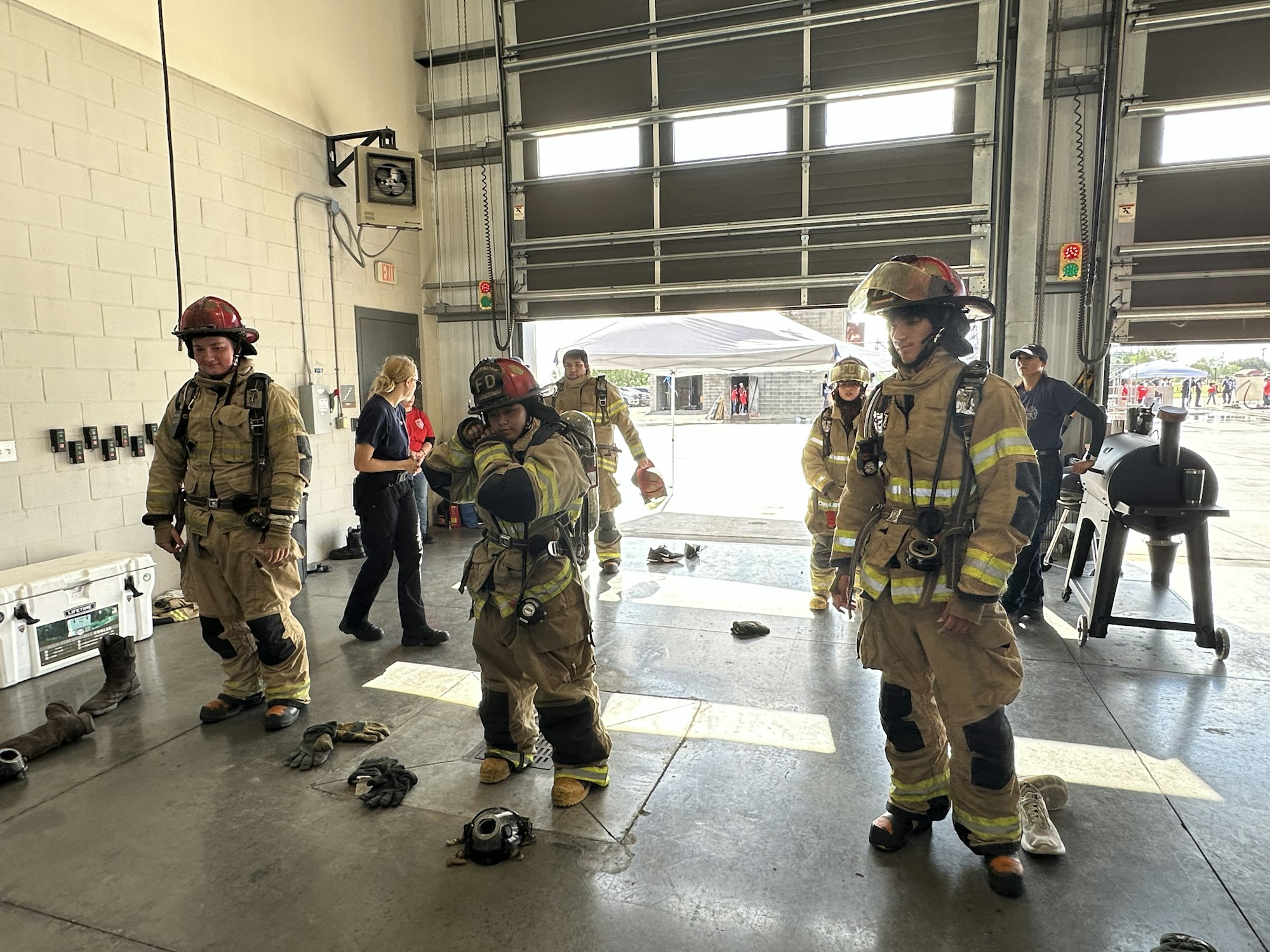 Firefighters in gear inside a garage, some equipment on the floor, people in casual clothes observe.