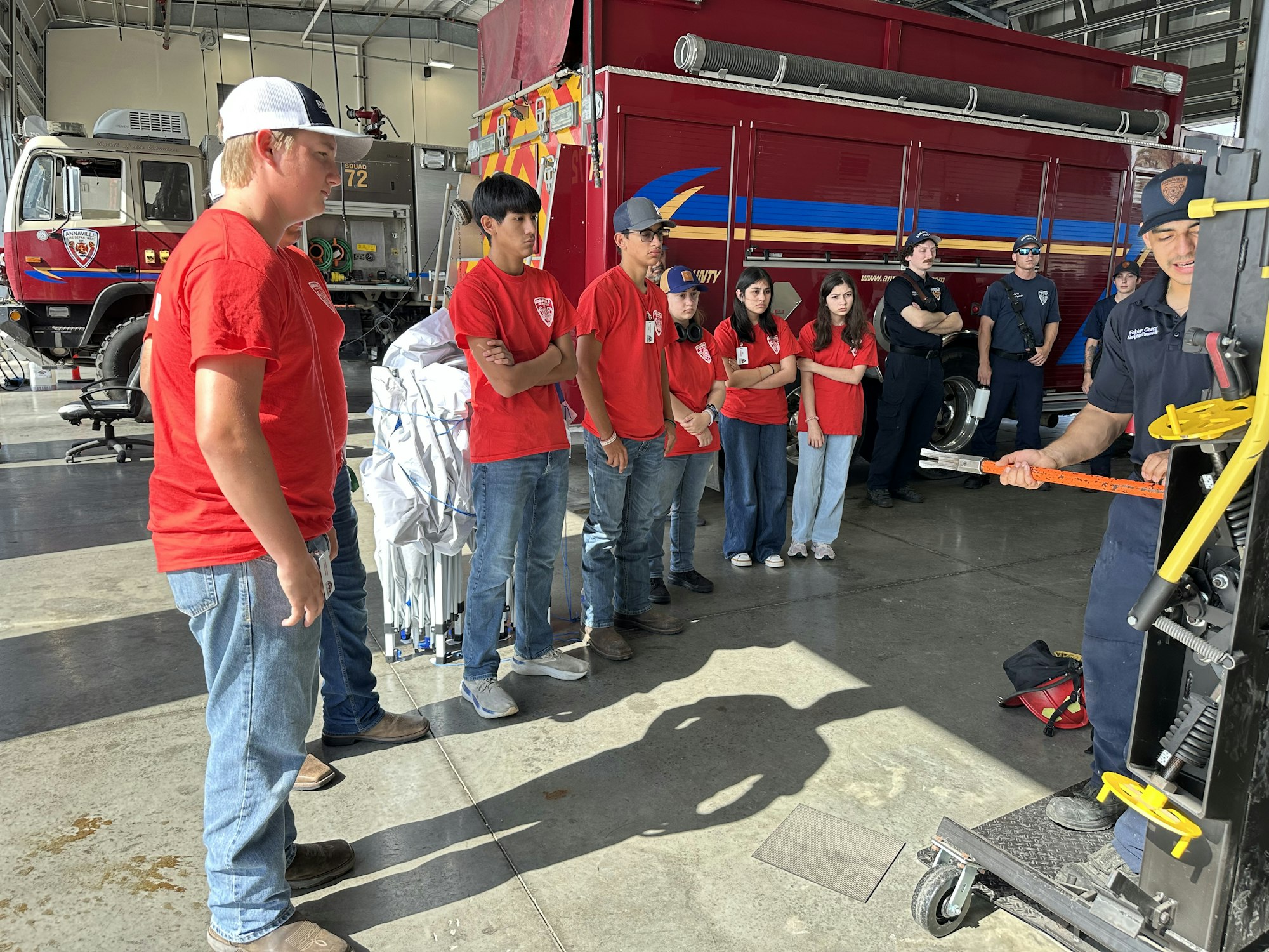A group of people in red shirts and firefighters watching a demonstration in a fire station.