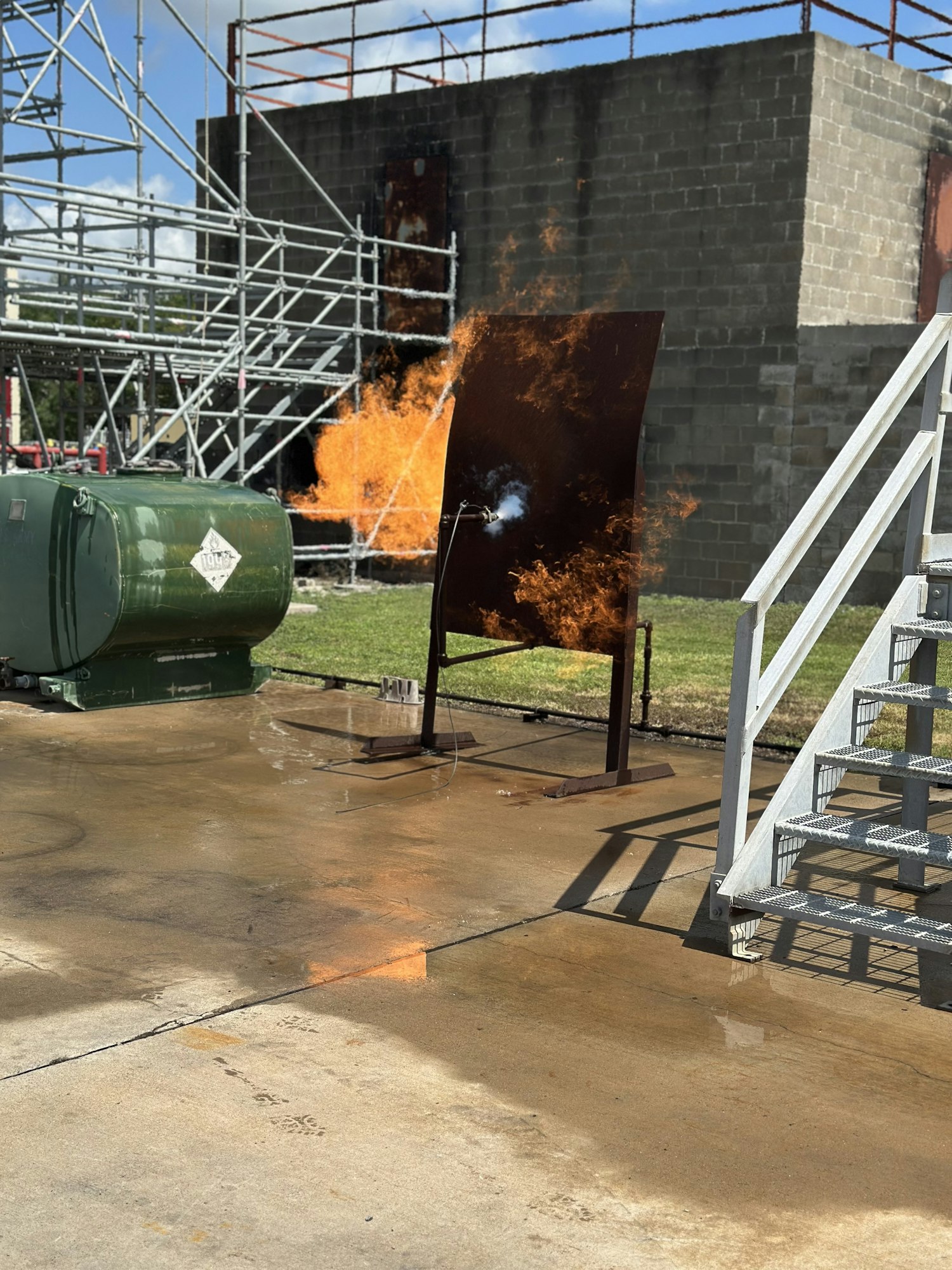A fire test with a sheet of metal and flames, scaffolding, and a caution-marked tank in an industrial setting.