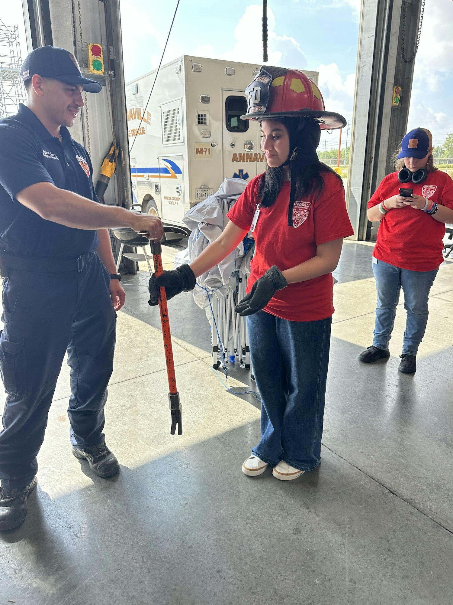 A person in a fire helmet holds a tool, guided by a firefighter. Another person in red watches nearby. An ambulance is in the background.