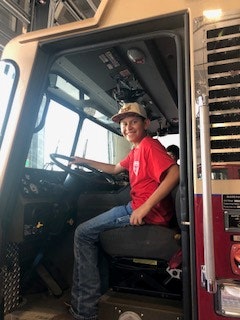 A person wearing a red shirt and hat is sitting in the driver's seat of a fire truck.