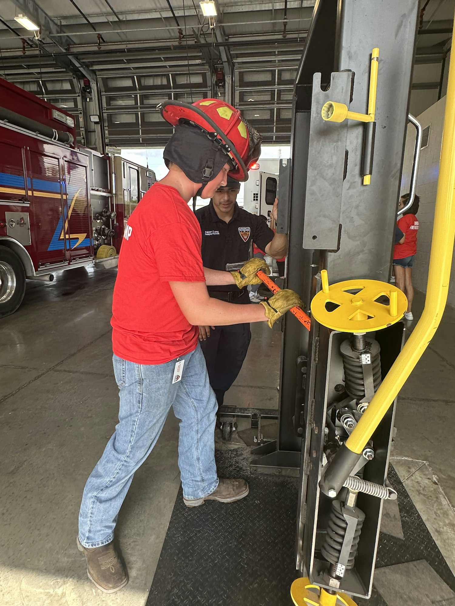 Two firefighters are inspecting or working on machinery inside a fire station, with a fire truck visible.