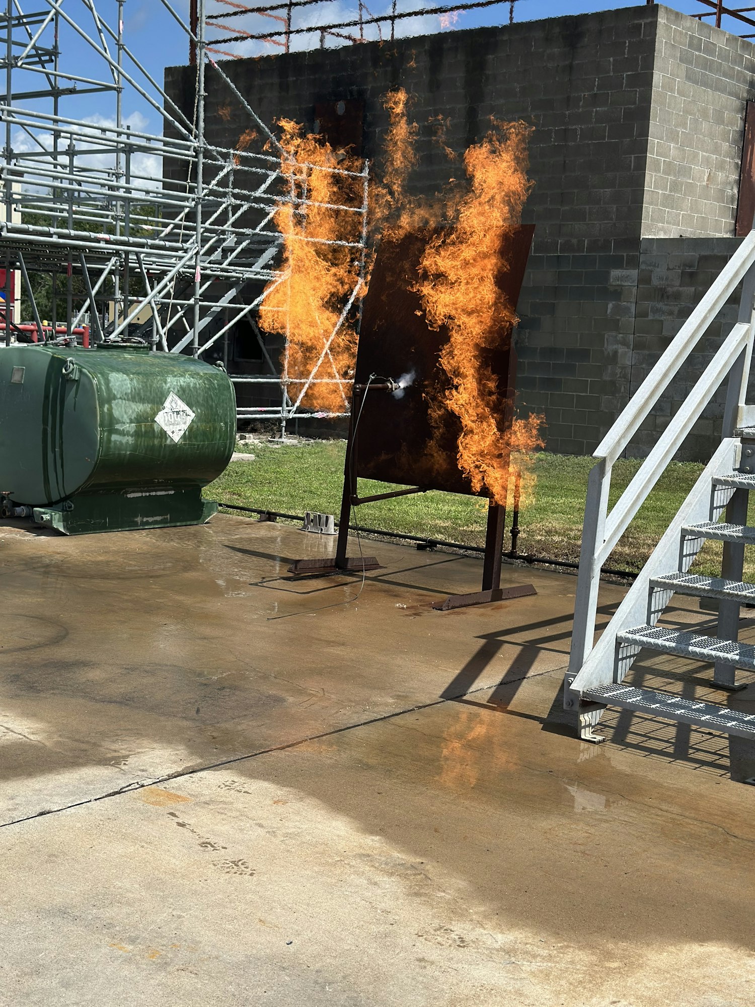 Fire blazing from a panel in a training area, with metal scaffolding and a nearby cylindrical tank on a concrete surface.