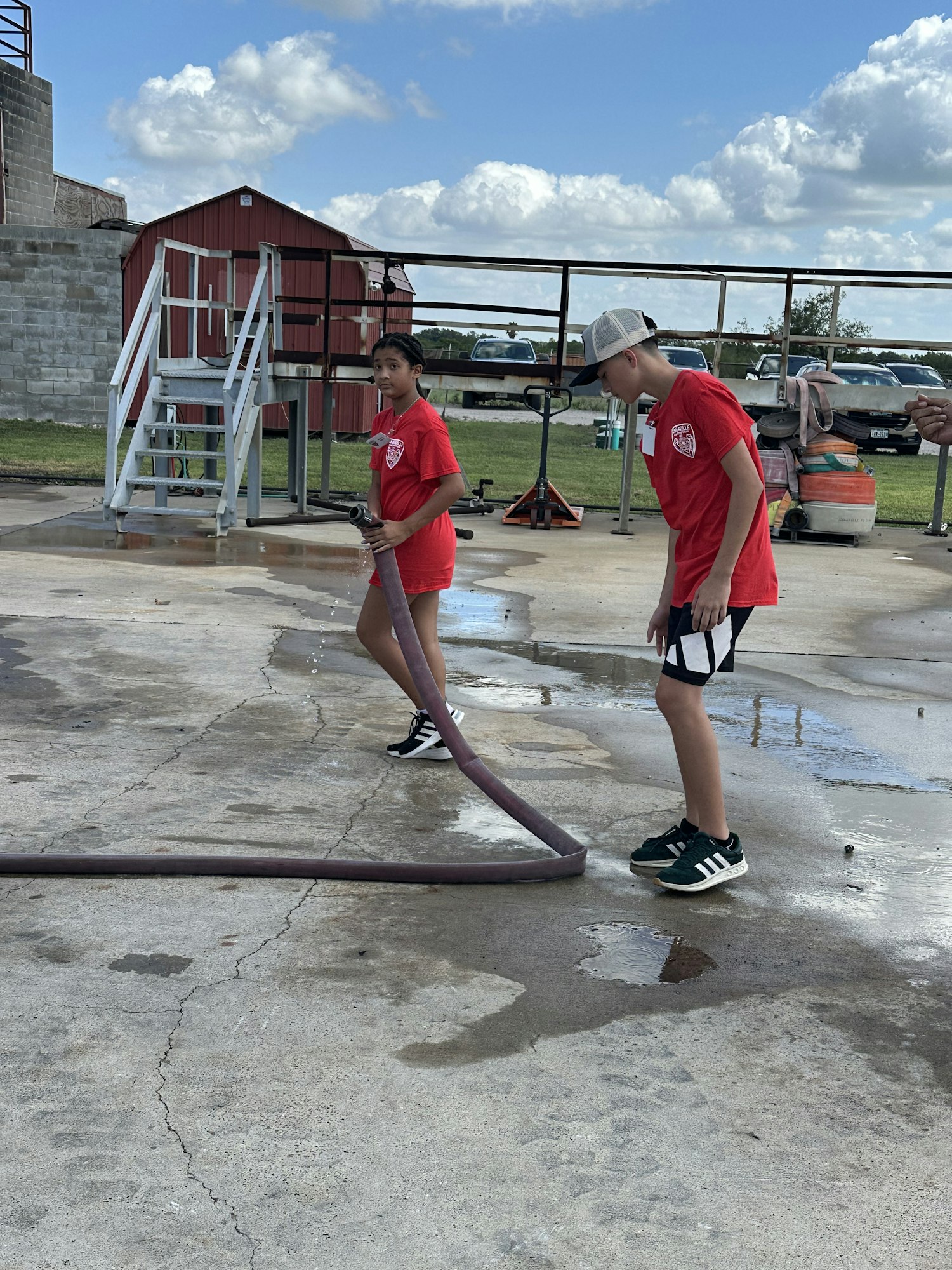 Two people in red shirts are handling a hose outdoors on a concrete surface near a barn, with cars in the background.