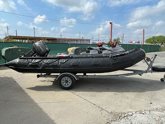 An inflatable black boat with an outboard motor sits on a trailer in a concrete lot under a cloudy sky.