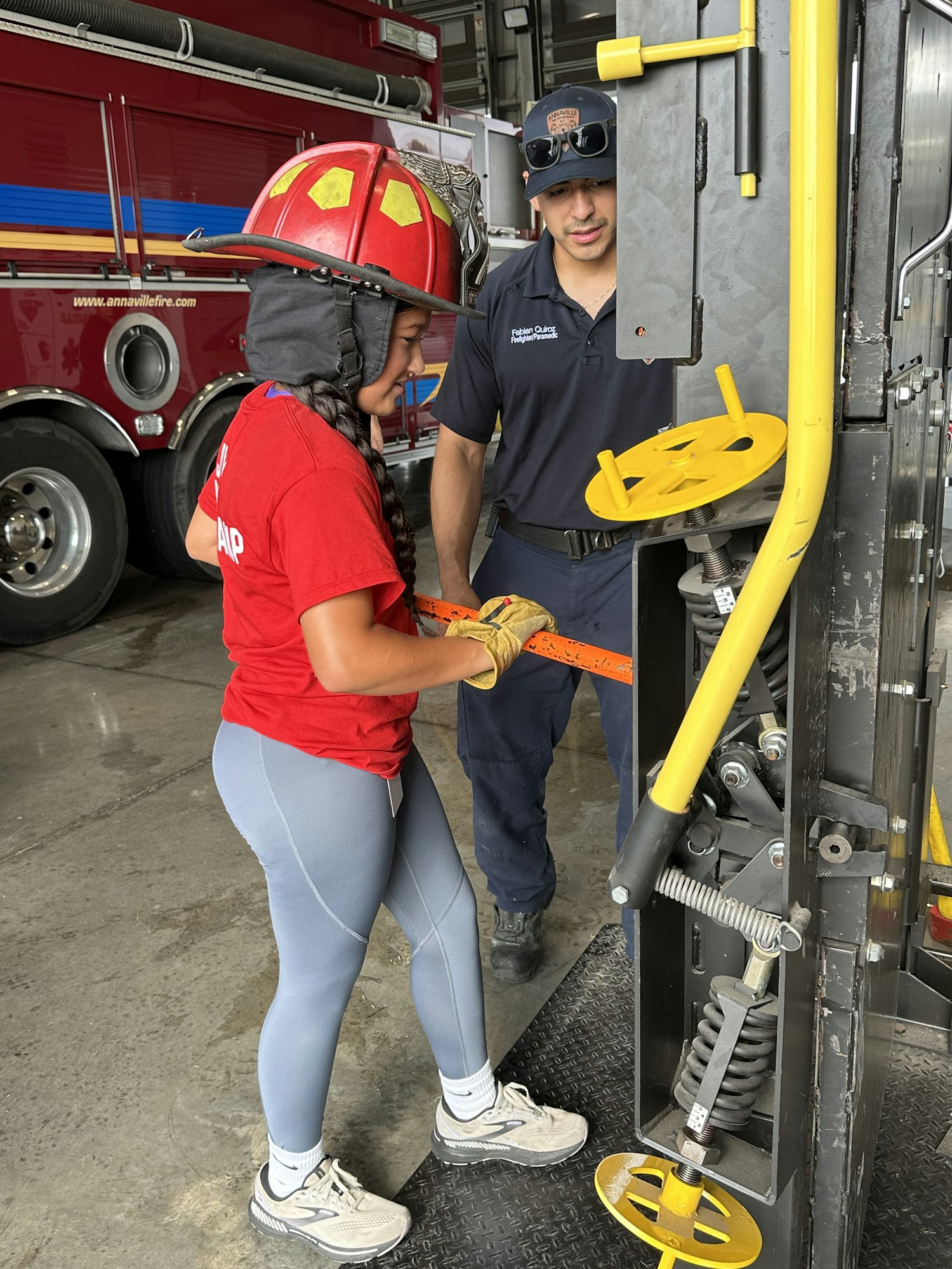 A person in a red helmet operates equipment, assisted by another person in a cap; a fire truck is in the background.