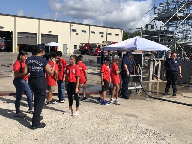 A group of people in red shirts and firefighters are gathered outdoors near a fire station.