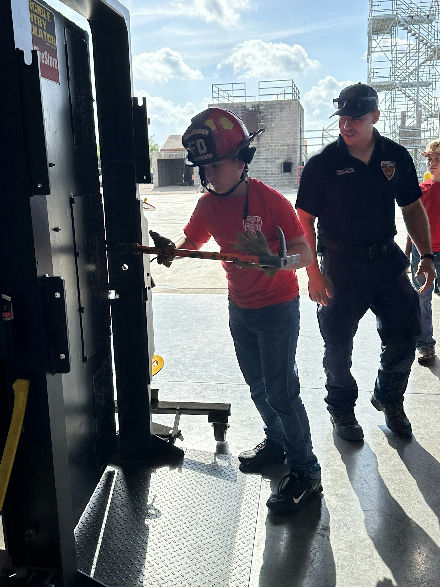 Two people, one in a red helmet, are practicing using a tool on a metal door simulator.