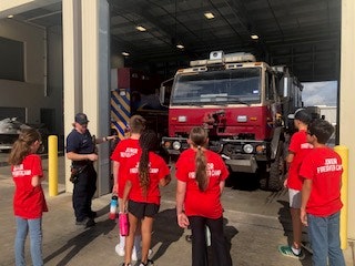A group of people in red "Junior Firefighter Camp" shirts standing in front of a fire truck inside a garage.
